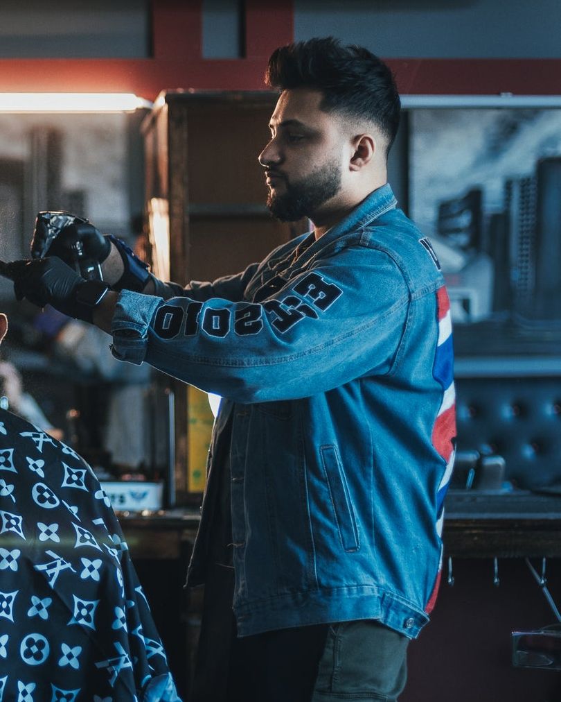 A barber in a denim jacket spraying water onto a seated client's head, mid-cut.