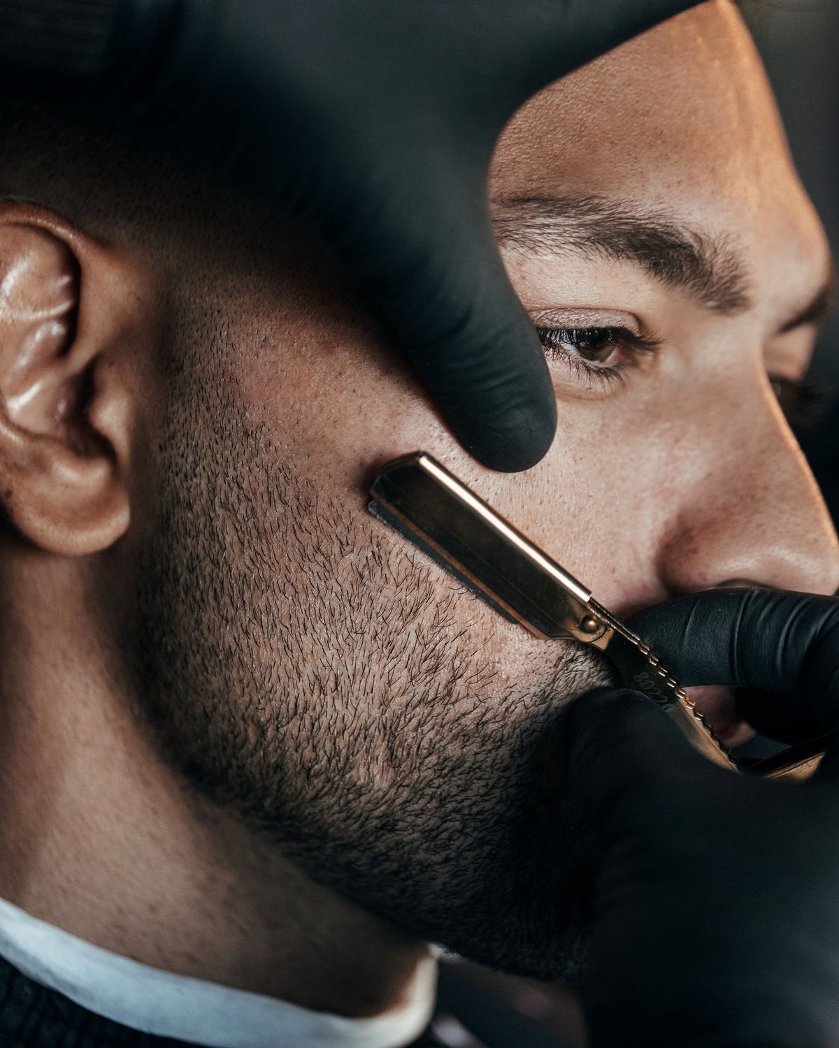 A barber steadying a client's cheek with one gloved hand while drawing a straight razor along his jawline.
