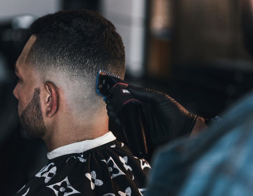 A barber in a denim jacket running clippers up the back of a client's head, taking a fade down to the skin.